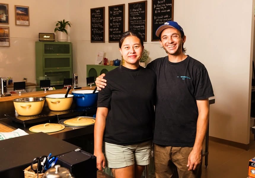 Two smiling people standing at the Samila Gaeng counter beside pots of curry and chalkboard menus