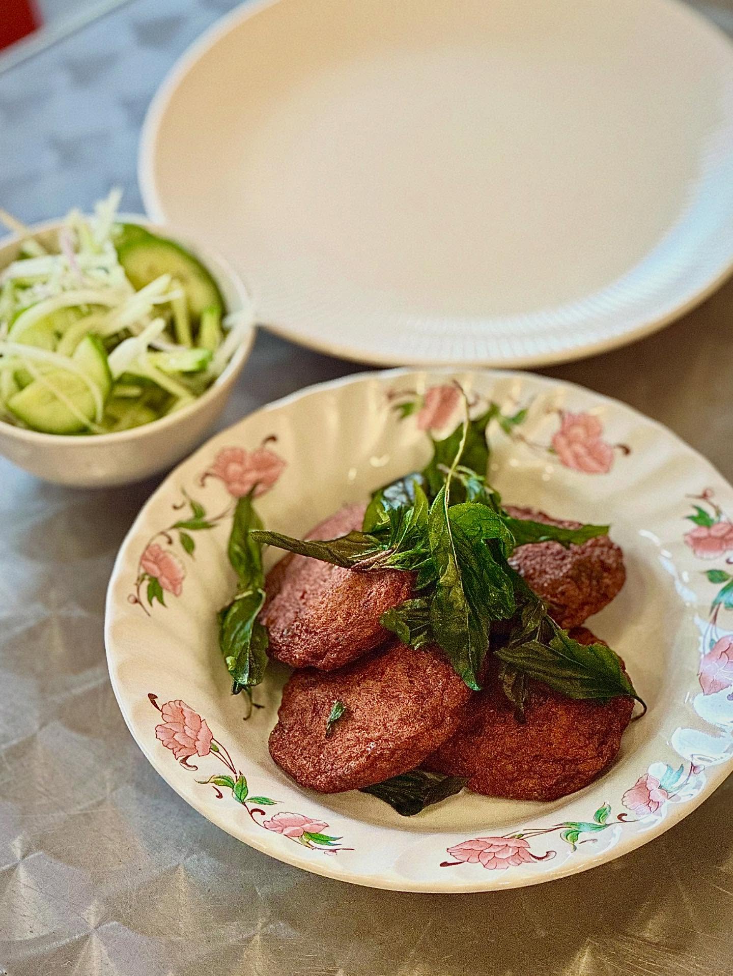 Fried fish cakes topped with crisp basil leaves on a floral plate, with a cucumber salad beside it